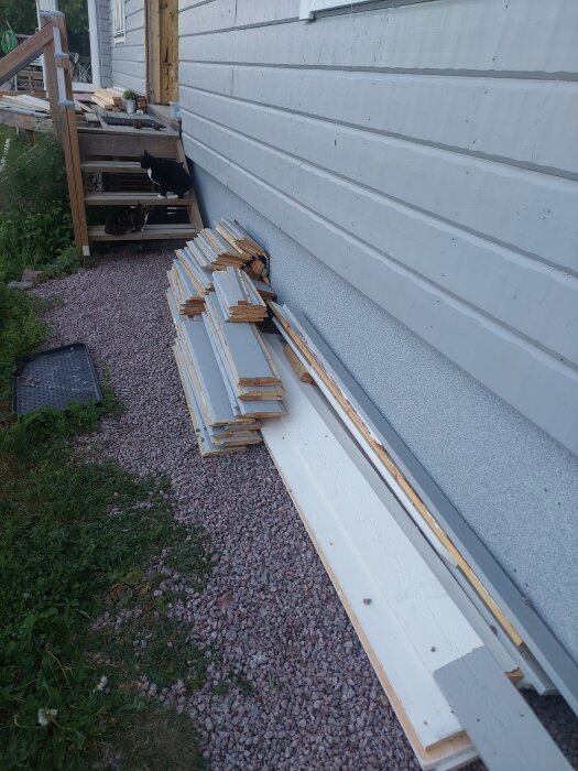 Staplat virke vid en husvägg, bredvid en trappa med två katter, en svart och en flerfärgad, sittande på trappstegen.
