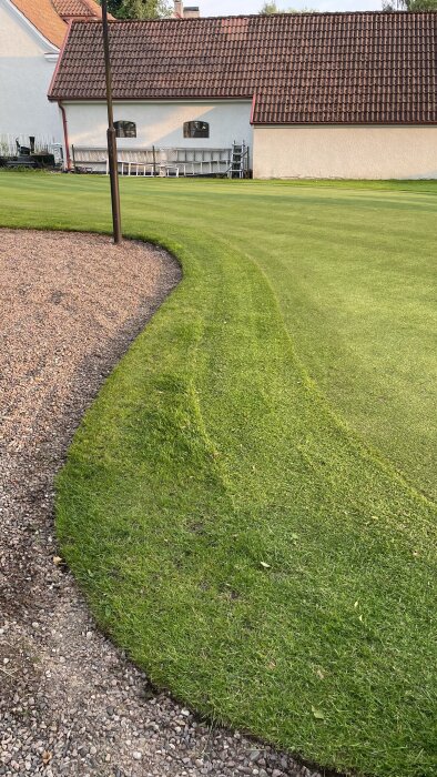 En välskött green med kantskuren gräsmatta vid grusgång, intill ett hus med tegelrött tak.