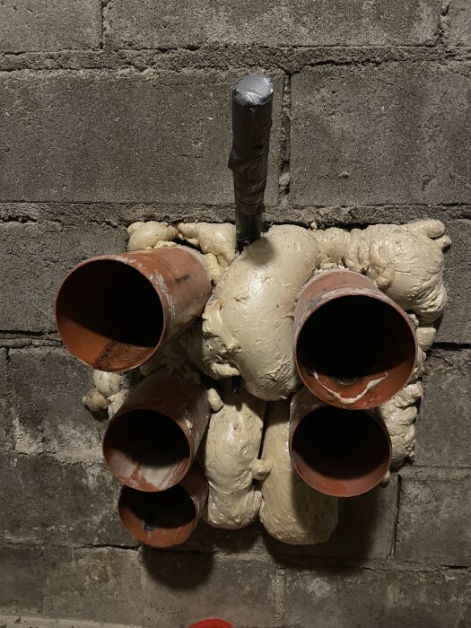 Fem bruna rör och en elkabel inkapslade i fogskum på en källarvägg av betongblock.