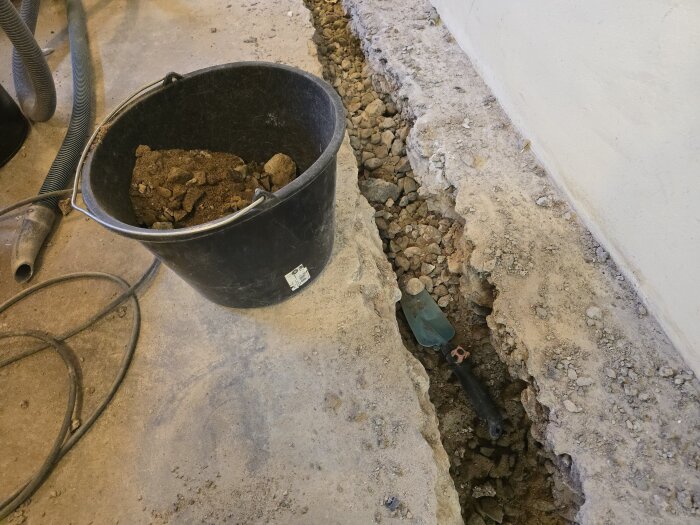 En svart hink full med sand och sten står bredvid en lång, smal utgrävd gång i ett betonggolv med synliga vattenrör.