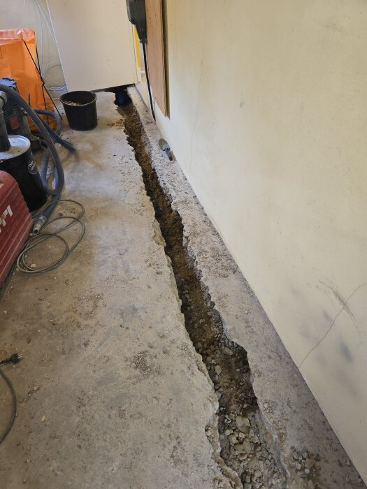 En smal ränna i ett betonggolv med grus och sten, verktyg och utrustning nära, del av ett renoveringsprojekt för avloppsinstallation.