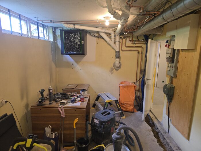 Källarrum under renovering med synliga rör och kablar i taket, arbetsyta fylld med verktyg, öppen dörr och påbörjad golvutgrävning.