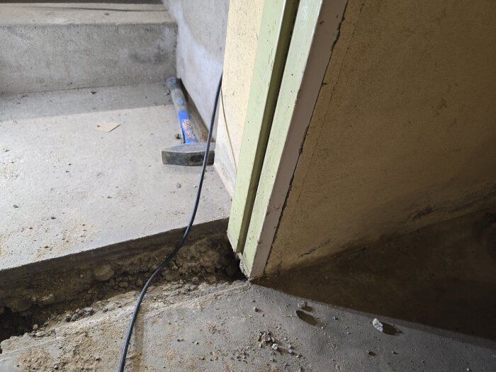 Betonggolv med avtäckt ränna och kabel, bredvid en delvis demonterad dörrkarm i en källare, med ett liggande verktyg.