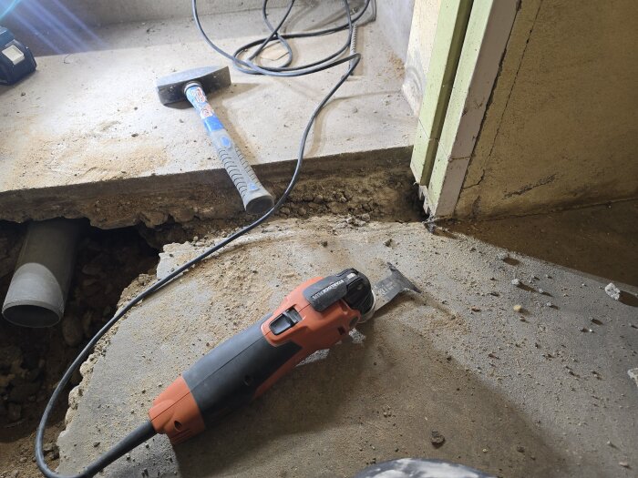 Multimaskin på betonggolv nära dörrkarm, synliga kablar och rör. Förberedelse för bygge och justering av avloppsrör i hobbyrum.