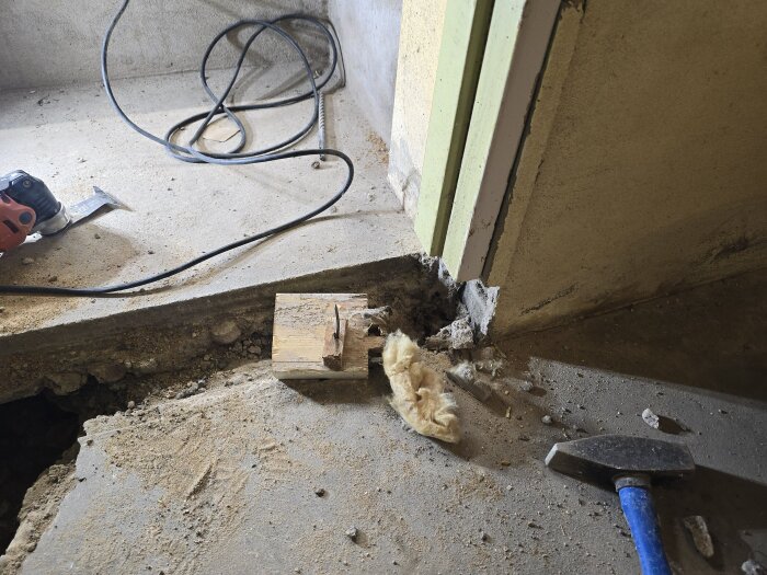Bilden visar en utgrävd ränna i cementgolv med verktyg och isoleringsmaterial bredvid. En dörrkarm med trasigt hörn är synlig i bakgrunden.