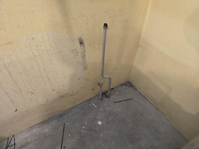 Ett hörn av ett rum med en gul vägg, ett uppstickande värmerör och en betonggolvyta med småskräp.