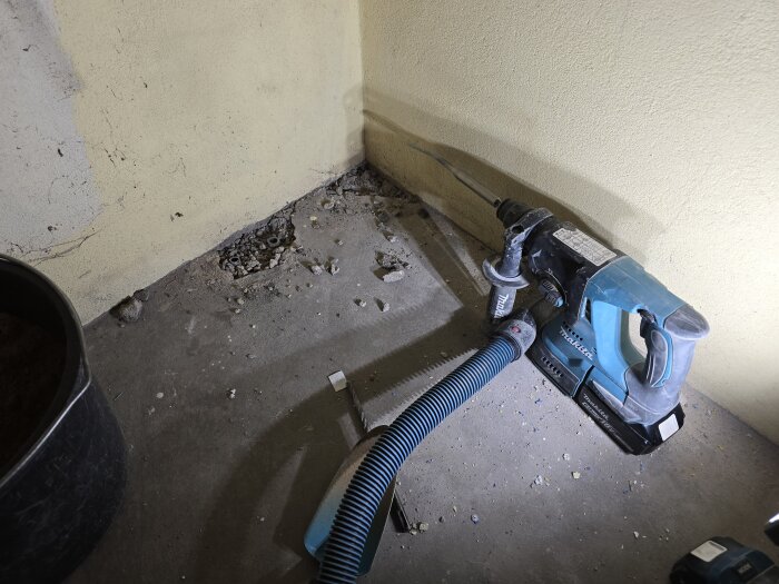 Hörnet av ett rum med borrhammare på golvet, intill en vägg som visar tecken på utgrävning och betongbitar spridda på golvet.