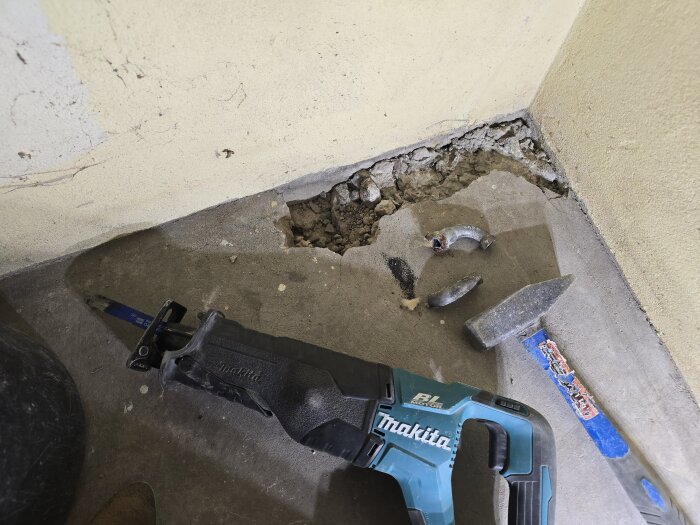 Hål i betonggolv med verktyg, en blå Makita såg och hammare ligger bredvid, samt böjda metallrörsdelar på golvet i ett hörnrum.