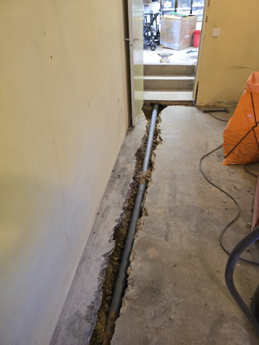 Ränna med delvis synligt avloppsrör grävd i betonggolv, vägg till vänster och dörr i fonden, rummet har pågående renoveringsarbete.