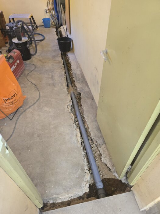 Ränna i golvet med nytt avloppsrör installerat, omringat av betong och sand; renoveringsutrustning synlig i bakgrunden.