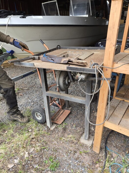 Person använder cirkelsåg på stålställning utomhus, med båt och trästruktur i bakgrunden. Flisig mark syns under arbetsområdet.