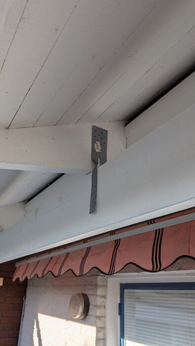 Metallfäste monterat på en träbalk under ett tak med synliga plankor. Det finns en markis i bakgrunden och en lampa på väggen.