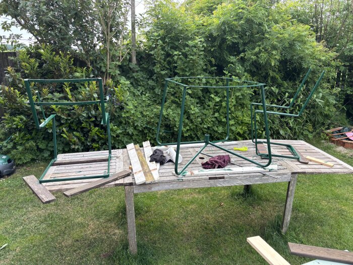 Trädgårdsmöbler på ett bord för renovering, med grönmålade stålramar, omgivna av buskar och träplankor.