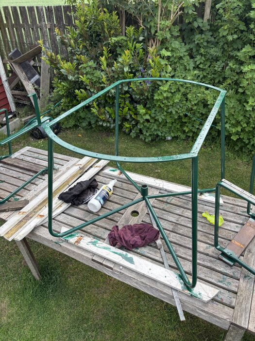 Gröna trädgårdsstolramar på ett träbord, med renoveringsprodukter samt trasa, omgivet av grönska och staket i bakgrunden.