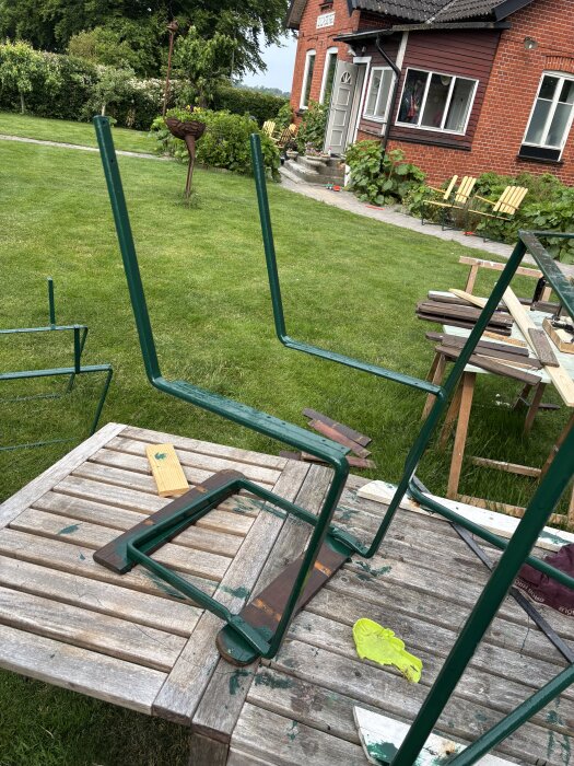 Gröna metallstolramar upp och ner på ett trädgårdsbord, med en röd tegelbyggnad i bakgrunden. Trädgårdsmiljö med gräsmatta och buskar.