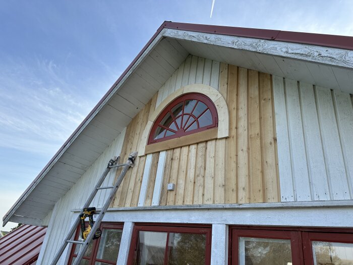 Halvmånefönster monterat på ett hus med röd ram, stege och borrmaskin synliga, del av pågående renoveringsarbete.