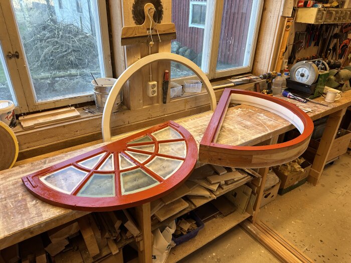 Halvmåneformat fönster på arbetsbänk, ommålat med röda ramar, i verkstadsmiljö med träverktyg och material i bakgrunden.