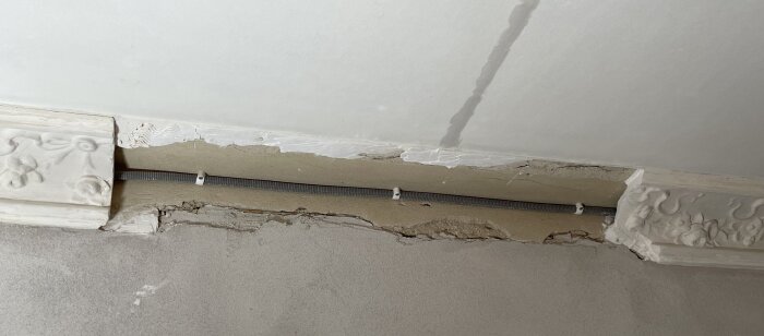 Skadad vägg med sprickor och exponerad kabel i taket, kring dekorerad gipspanel.