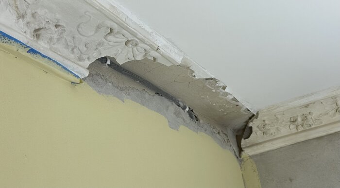 Sprucken taklist med synliga ledningar, gult och grått väggområde, del av renoveringsprocess.