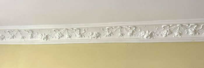 Vit dekorerad taklist med blommotiv mot gul vägg.