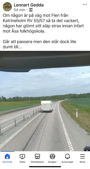 En släpvagn står på vägen bredvid en asfalterad körbana omgiven av gröna fält och skog, fotograferad genom en bilruta.