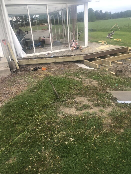 Bild på ett halvfärdigt trädäck vid en inglasad veranda med utemöbler och leksaker. Ett stuprör och spade syns vid den öppna ytan.