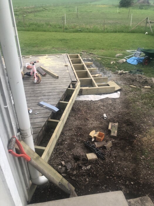 Byggnation av trätrall pågår vid husvägg med stuprör, spade lutad mot väggen, och barncykel på trallen. Grus och material på marken.