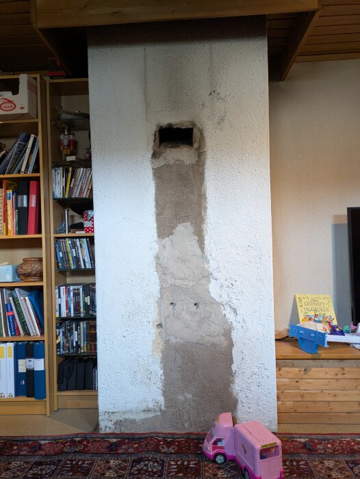 Murstock i ett 70-tals hus, delvis avlägsnat för att installera en ny kamin, med sliten yta och ett rosa leksaksfordon i förgrunden.