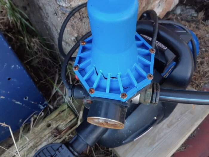 Blå tryckregulator med rostiga skruvar monterad på en pumpenhet utomhus, omgiven av kablar och vegetation.