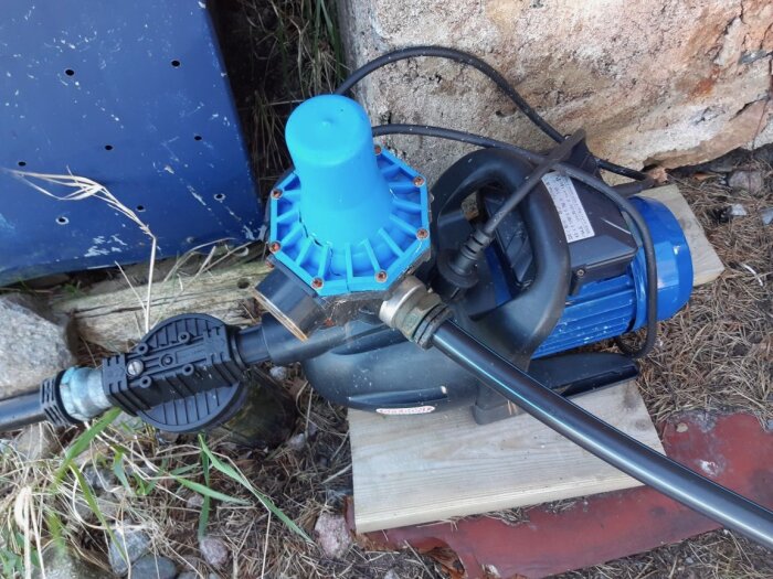 Saltvattenpump monterad utomhus, stående på en träplatta och skyddad av en blå kåpa. Synliga komponenter och kopplingar, omgiven av sten och växtlighet.