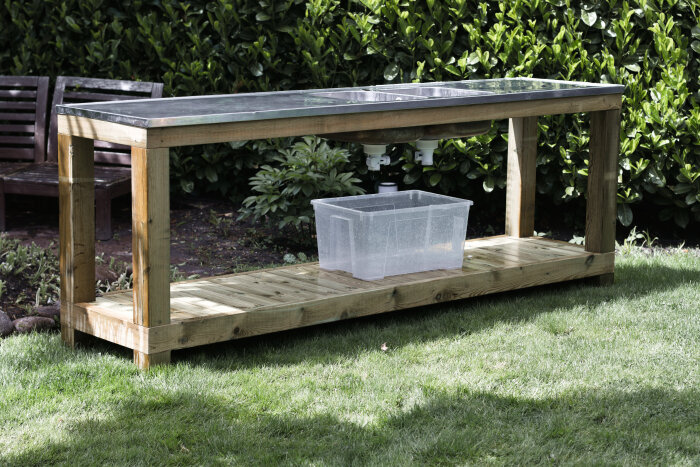 Utomhuskök av trä med rostfri diskho och plastlåda under, placerat på en gräsmatta med buskar i bakgrunden.