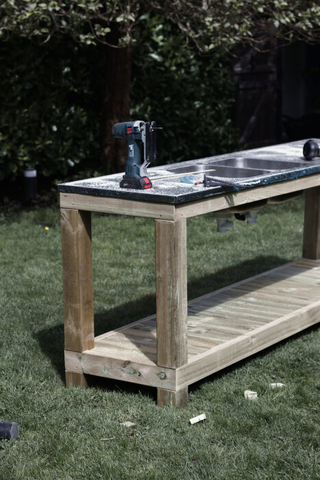 Utomhus bänkskiva av trä med en rostfri diskho och en hylla under, placerad på gräsmatta med en elektrisk såg ovanpå.