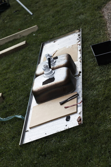 Rostfri diskbänk med dubbelho och kranar på marken, omgiven av verktyg och byggmaterial, del av ett omplanteringsbänkprojekt.