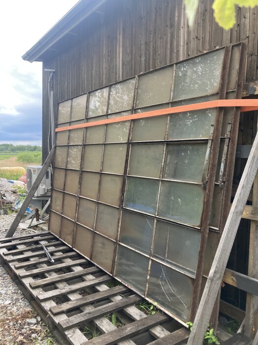 Gamla fönsterpaneler lutade mot en träbyggnad, med spruckna och smutsiga rutor, på en sliten träpall utomhus.