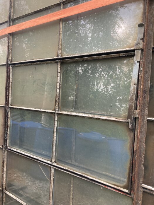 Gammalt industrifönster med slitet glas och rostiga metallramar, med spegling av blåa tunnor utanför.
