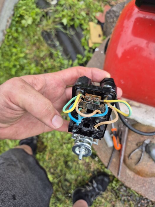 En person håller i en tryckvakt med vatten- och rostskador. Bakgrunden visar gräs och verktyg.