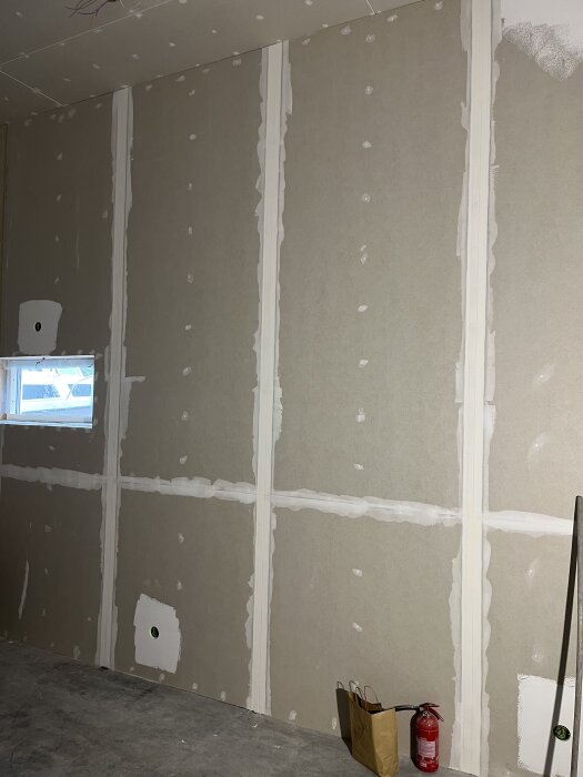 Väggar med pågående spacklingsarbete, synliga skarvar och spackelfläckar. Ett fönster, brandsläckare och påse syns i rummet.