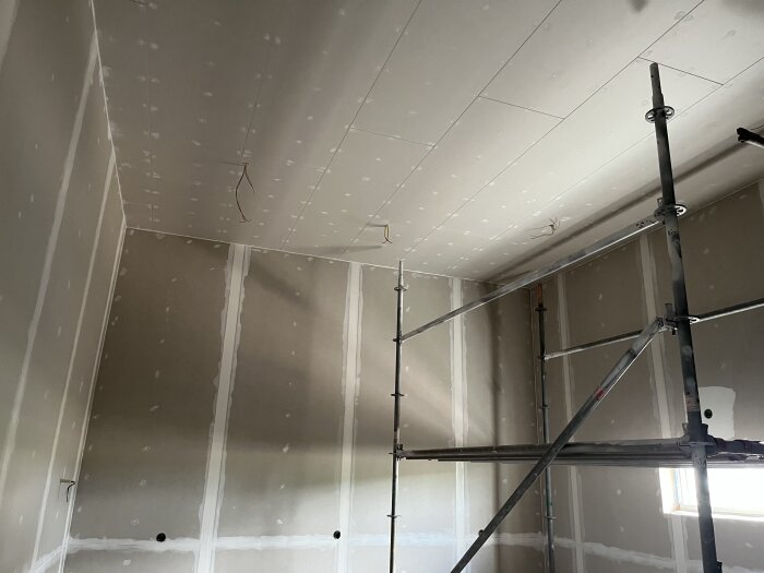 Rum under renovering med spacklade väggar och tak, stege i hörnet. Kablar hänger från taket där belysning har testats.