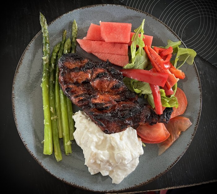 Grillad fläskkarré med potatissallad, vitlöksfrästa sparrisar, vattenmelon och grönsaker på en tallrik.