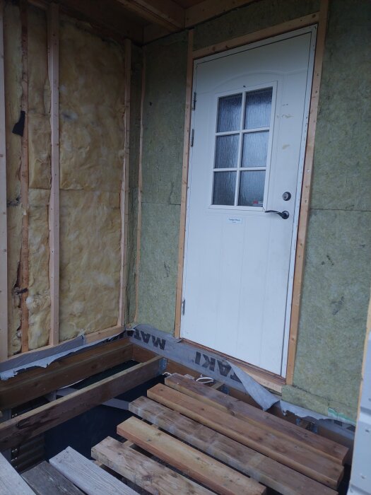 Väggstruktur under renovering med isolering synlig, träreglar och en vit dörr med glasrutor, samt brädor framför dörröppningen.