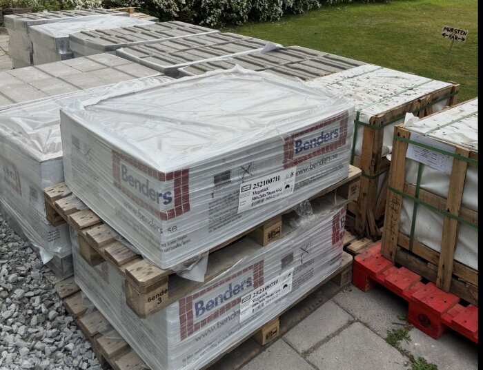 Pall med inplastad mursten från Benders på en stengång, omgiven av grönska. En skylt pekar mot "Mursten här".