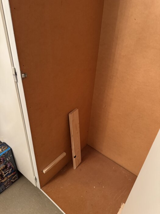 Ett öppet skåp från 60-70-talet, visar insidan med tunn MDF-skiva. En vertikal bräda och hyllstöd finns nära botten av skåpet.