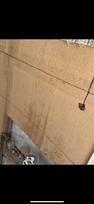 Brun skiva monterad på väggen med flera synliga skarvar och hål från skruvar. Undre kant nära rör och ventil, del av byggprojekt.