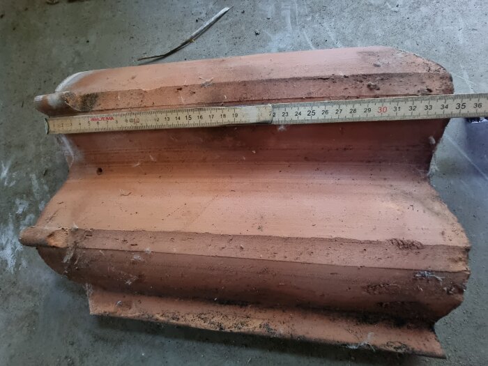 Tvåkupig takpanna med en tumstock placerad ovanpå för att mäta längd; pannans yta är dammig och något sliten.