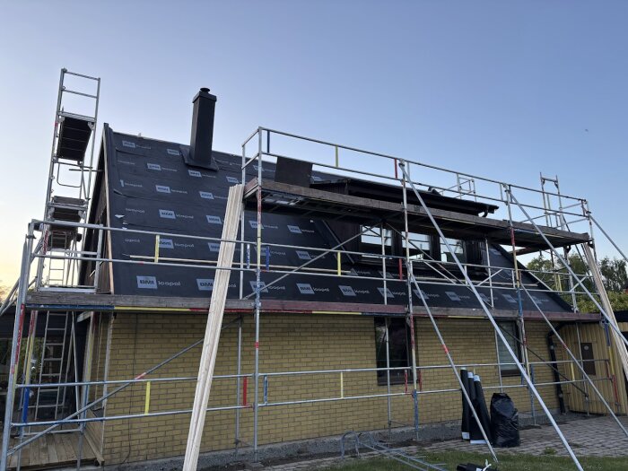 Hus under takrenovering med byggställning, ny takpapp synlig och plåtdetaljer påbörjade, blå himmel i bakgrunden.