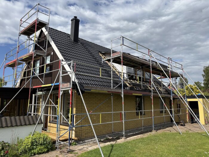 En villa med svart tak under renovering, omgiven av byggställningar. Det nya taket är delvis klart och arbetsmaterial syns bredvid huset.