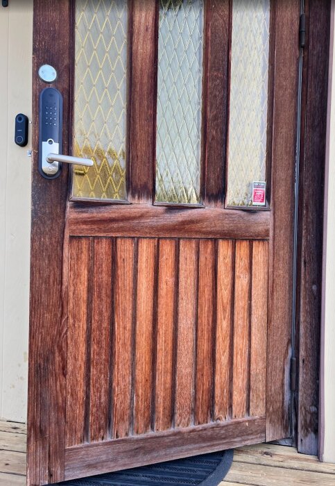 Trädörr med glasdetaljer och ett digitalt dörrlås, synliga sprickor i träet längst ner till höger.