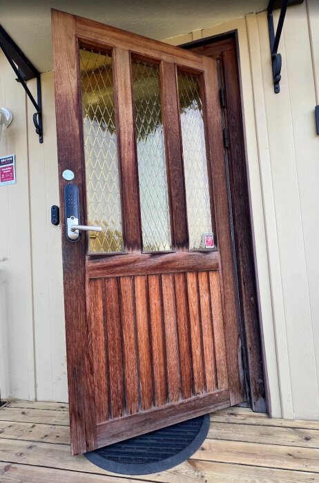 Träytterdörr med glaspanel och synliga sprickor, står halvöppen mot beige vägg. Dörren har metallhandtag och står på trätrall.