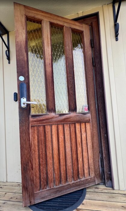 Trädörr med glasrutor, några sprickor synliga. Dörren är utrustad med ett digitalt lås och ståröppen, redo för renovering.