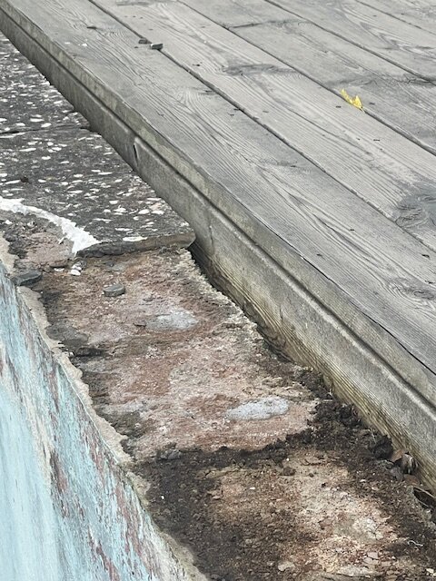 Nedslitna stenplattor kring en gammal poolkant, med ett intilliggande trädäck. Plattornas skick visar behov av renovering och trallen är i trä.